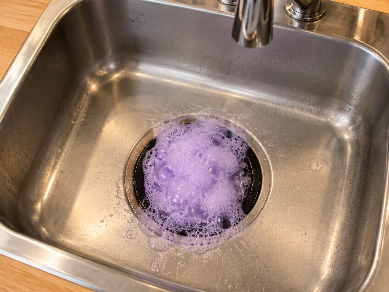 Purple foam bubbling up from a garbage disposal in a stainless steel kitchen sink